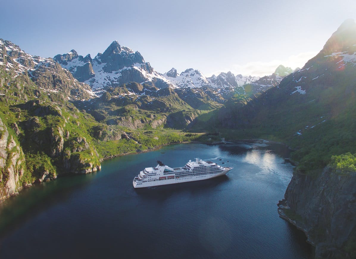 Cruise ship in a calm fjord surrounded by steep green cliffs and snow-capped mountains under a clear sky.