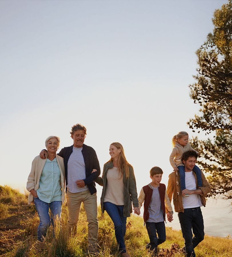 A family of six, including a child on shoulders, walks through a grassy field at sunset, smiling and enjoying the outdoors.