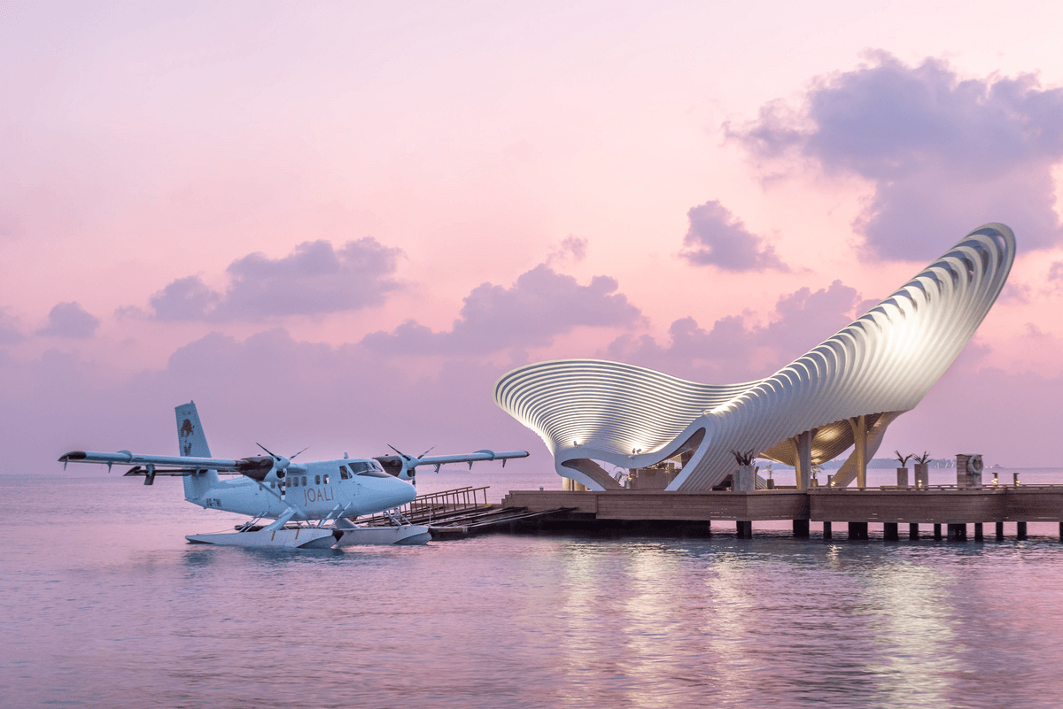 A seaplane docked near a futuristic, shell-like terminal against a pink sunset sky and calm water.
