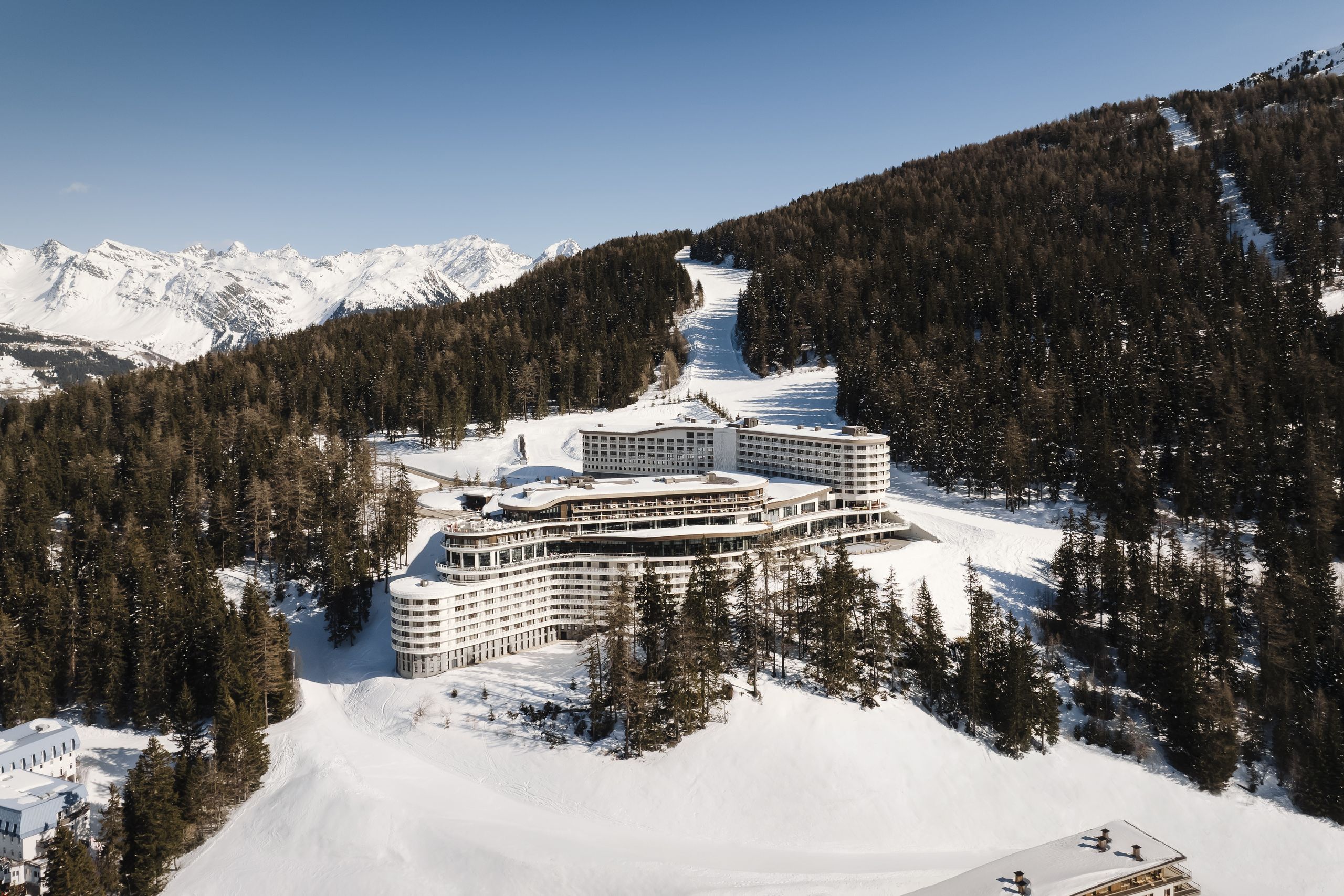 A large, modern hotel nestled in a snowy mountainous landscape, surrounded by dense pine forests and ski slopes under a clear blue sky.